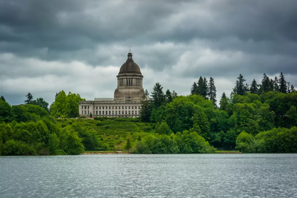 View of Washington state capitol building dome from across lake with green trees in foreground and grey cloudy sky
